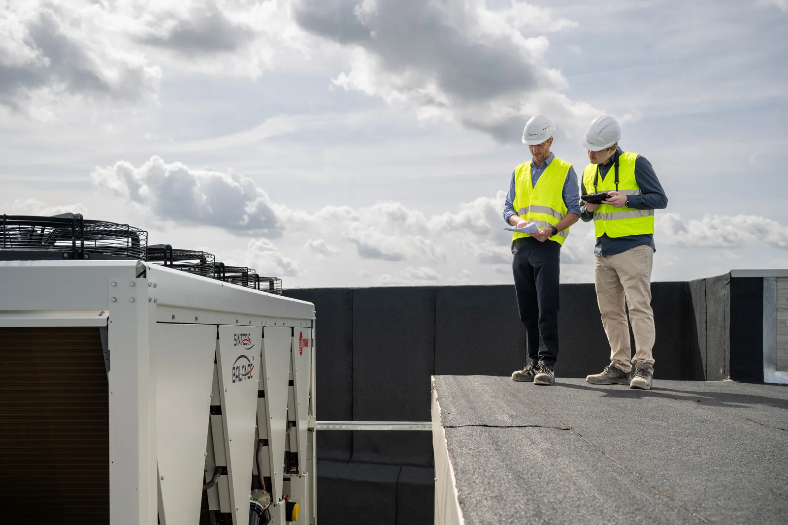 Twee ingenieurs op het dak bij de klimaatgroep: de een met het plan in de hand, de ander met de tablet, tegen een wolkenlucht.