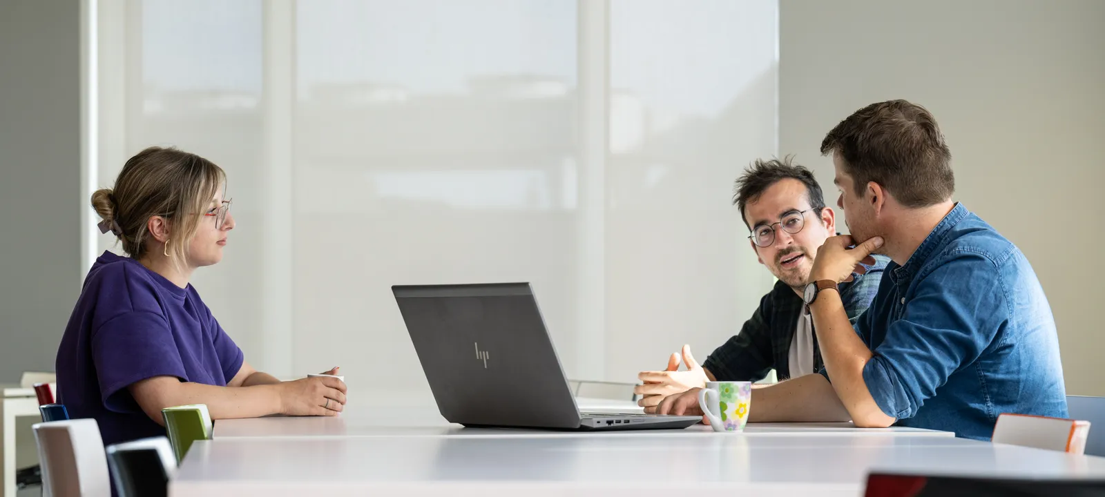 Drie collega's in de vergaderzaal rond een laptop: een vrouw in het paars tegenover twee mannen in gesprek.