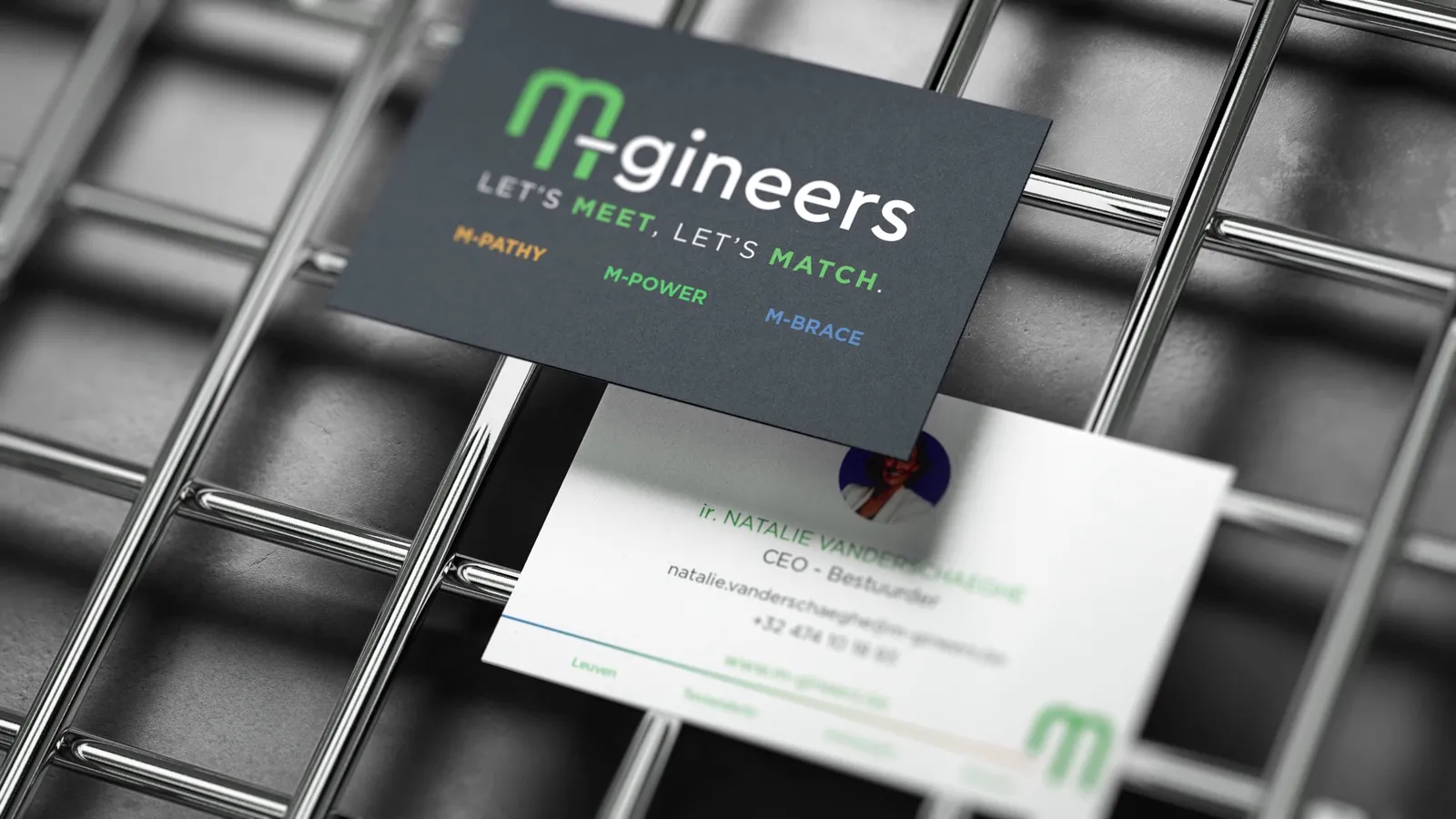 Twee visitekaartjes op een metalen rooster: het donkere kaartje met het logo en 'Let's meet, let's match.' boven M-pathy, M-power en M-brace, daaronder het witte kaartje met de contactgegevens.