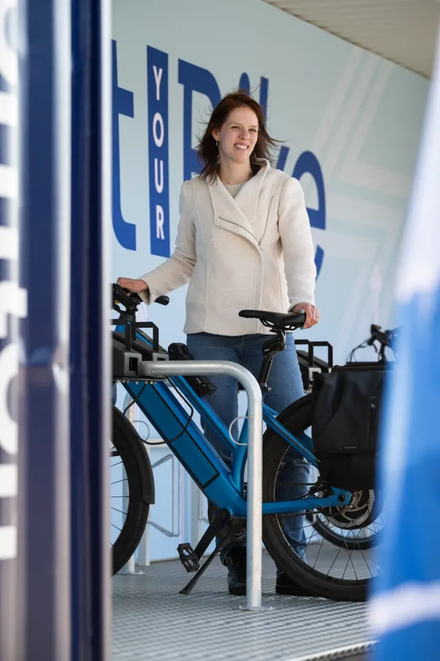 Een vrouw in een lichte wollen jas staat glimlachend bij een blauwe e-bike in de fietscontainer, voor een wand met grote blauwe letters.